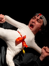 Scena z przedstawienia: mężczyzna trzymający lalkę ubraną w biały, sportowy strój, w miejscu głowy lalki widać głowę aktora. Poza lalki to poza karate z jedną nogą uniesioną do góry.