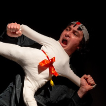 Scena z przedstawienia: mężczyzna trzymający lalkę ubraną w biały, sportowy strój, w miejscu głowy lalki widać głowę aktora. Poza lalki to poza karate z jedną nogą uniesioną do góry.