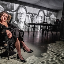 Kobiera w czarnej sukni siedzi na fotelu, za nią czarno-białe fotografie, stół i postać stojąca w cieniu.