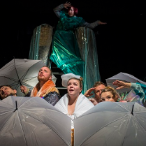 Kilka osób za rozłożonymi parasolami, patrzą w górę. Za nimi kobieta w zielonym połyskującym stroju.