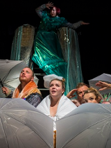 Kilka osób za rozłożonymi parasolami, patrzą w górę. Za nimi kobieta w zielonym połyskującym stroju.