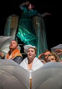 Kilka osób za rozłożonymi parasolami, patrzą w górę. Za nimi kobieta w zielonym połyskującym stroju.