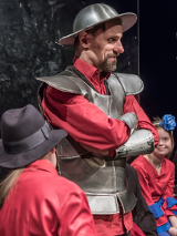 Mężczyzna w blaszanym kapeluszu - hełmie na głowie i stroju przypominającym zbroję, pod którą ma czerwoną koszulę. Ma ręce założone na piersi i delikatnie uśmiecha się.