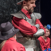 Mężczyzna w blaszanym kapeluszu - hełmie na głowie i stroju przypominającym zbroję, pod którą ma czerwoną koszulę. Ma ręce założone na piersi i delikatnie uśmiecha się.