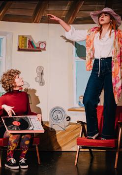 Dwie młode dziewczyny, jedna siedzi i trzyma na kolanach płyę winylową, druga stoi z rozłożonymi rękoma na fotelu.