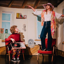 Dwie młode dziewczyny, jedna siedzi i trzyma na kolanach płyę winylową, druga stoi z rozłożonymi rękoma na fotelu.