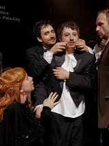 Dwóch mężczyzn soi blisko trzeciego mężczyzny, jeden z nich dotyka jego twarzy. Dwie kobiety przygladają się mężczyznom.