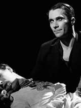 Czarno-białe zdjęcie, na którym jeden mężczyzna podtrzymuje drugiego mężczyznę. Ciemne tło, na drugim planie czterokołowy wózek kelnerski.