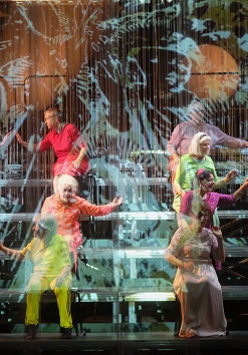 Na zdjęcie kilkanaście osób w kolorowych strojach, siedzą na stopniach w czterech rzędach, zwróceni parami do siebie, niektórzy z wyciągniętymi rękoma.