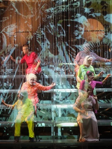 Na zdjęcie kilkanaście osób w kolorowych strojach, siedzą na stopniach w czterech rzędach, zwróceni parami do siebie, niektórzy z wyciągniętymi rękoma.