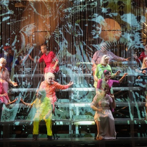 Na zdjęcie kilkanaście osób w kolorowych strojach, siedzą na stopniach w czterech rzędach, zwróceni parami do siebie, niektórzy z wyciągniętymi rękoma.