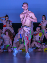 Grupa tancerzy na scenie. Na pierwszym planie mężczyzna z nagim torsem, w kolorowych spodniach i sportowych butach.