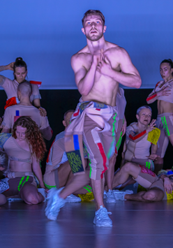 Grupa tancerzy na scenie. Na pierwszym planie mężczyzna z nagim torsem, w kolorowych spodniach i sportowych butach.