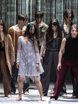 Grupa tancerzy w różnorodnych strojach, wszyscy stoją z opuszczonymi głowami.