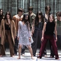 Grupa tancerzy w różnorodnych strojach, wszyscy stoją z opuszczonymi głowami.