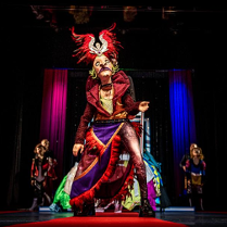 Na zdjęciu postać ubrana w kolorowy strój z maską na twarzy. Stoi na scenie na pierwszym planie, za nią w tle inne postacie.