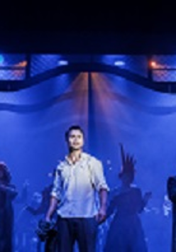 Zdjęcie w niebiesko-czarnych barwach: aktorzy na scenie, w centralnym pukcie mężczyzna w białej koszuli.