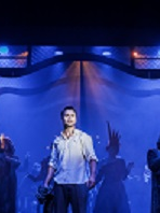 Zdjęcie w niebiesko-czarnych barwach: aktorzy na scenie, w centralnym pukcie mężczyzna w białej koszuli.