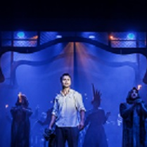 Zdjęcie w niebiesko-czarnych barwach: aktorzy na scenie, w centralnym pukcie mężczyzna w białej koszuli.