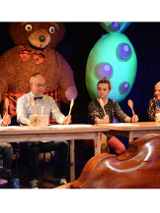 kadr ze spektaklu przedstawia czworo aktorów siedzących przy stołach i trzymających uniesione drewniane łyżki. Przed nimi leżą odwrócone do góry dnem garnki.