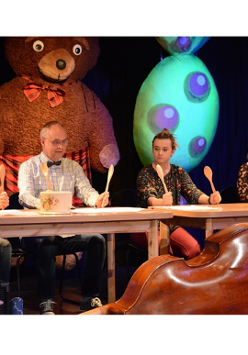 kadr ze spektaklu przedstawia czworo aktorów siedzących przy stołach i trzymających uniesione drewniane łyżki. Przed nimi leżą odwrócone do góry dnem garnki.