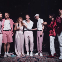 Siedmioro ludzi ubrani w odcienie różowego stoją na scenie wyłozonej dywanami. Za nimi instrumenty muzyczne.