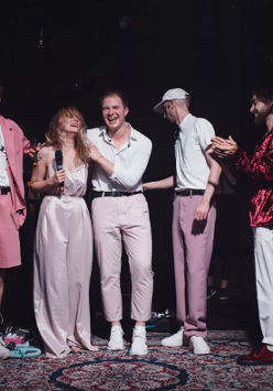 Siedmioro ludzi ubrani w odcienie różowego stoją na scenie wyłozonej dywanami. Za nimi instrumenty muzyczne.