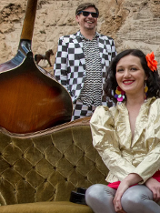 Kobieta w złotej marynarce siedzi na zielonej kanapie na plaży. Za nią mężczyna ubrany w garnitur w szachownicę. O kanapę oparty jest kontrabas. W tle kolorowy parawan w paski i ściana skalna.