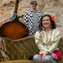 Kobieta w złotej marynarce siedzi na zielonej kanapie na plaży. Za nią mężczyna ubrany w garnitur w szachownicę. O kanapę oparty jest kontrabas. W tle kolorowy parawan w paski i ściana skalna.