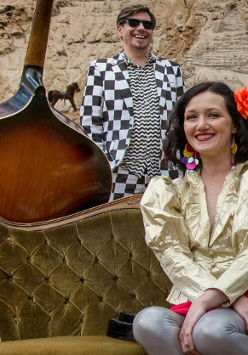 Kobieta w złotej marynarce siedzi na zielonej kanapie na plaży. Za nią mężczyna ubrany w garnitur w szachownicę. O kanapę oparty jest kontrabas. W tle kolorowy parawan w paski i ściana skalna.