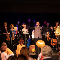 Zdjęcie przedstawia czlonków orkiestry, pośrodku dyrygent, wokół i za nim muzycy z instrumentami