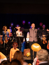 Zdjęcie przedstawia czlonków orkiestry, pośrodku dyrygent, wokół i za nim muzycy z instrumentami