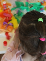 Zdjęcie zrobione z góry. Dziewczynka stoi tyłem do obiektywu. Przed nią na stoliku leżą kolorowe przybory plastyczne.