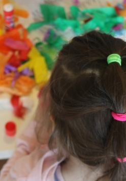 Zdjęcie zrobione z góry. Dziewczynka stoi tyłem do obiektywu. Przed nią na stoliku leżą kolorowe przybory plastyczne.