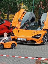 Dwa duże auta i jedno mniejsze, w którym siedzi mały chłopiec. W tle plac zabaw i drzewa.