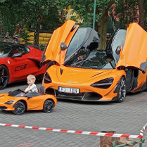 Dwa duże auta i jedno mniejsze, w którym siedzi mały chłopiec. W tle plac zabaw i drzewa.