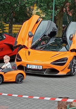 Dwa duże auta i jedno mniejsze, w którym siedzi mały chłopiec. W tle plac zabaw i drzewa.