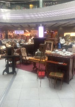 Różne przedmioty, w tym krzesła, stoły, stojaki oraz drewniane pianino w galerii handlowej.