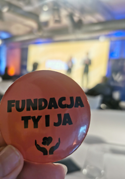 Dłoń trzymająca przypinkę z napisem "Fundacja ty i ja". Tło rozmazane.