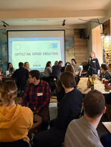 Grupa ludzi siedzi przy stołach w barze, uczestnicząc w wydarzeniu "speed debating". Na ścianie widać duży ekran z napisem "Witaj na speed debating'. Jedna osoba stoi z mikrofonem i przemawia do uczestników, a reszta rozmawia w małych grupach.