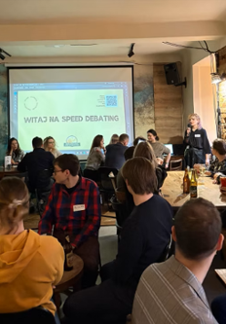 Grupa ludzi siedzi przy stołach w barze, uczestnicząc w wydarzeniu "speed debating". Na ścianie widać duży ekran z napisem "Witaj na speed debating'. Jedna osoba stoi z mikrofonem i przemawia do uczestników, a reszta rozmawia w małych grupach.