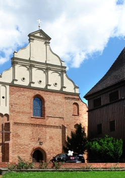 Zabytkowy kościół z cegły obok którego stoi drewniana dzwonnica. Fasada kościoła ma jasny szczyt. Dzwonnica ma ciemne, drewniane ściany. W tle widać niebo z białymi chmurami i drzewa otaczające budynki.