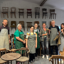 Grupa osób w fartuchach. Wokół przedmioty wykonane z drewna. W tle krzesła wiszące na szarej ścianie.