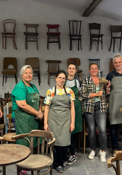 Grupa osób w fartuchach. Wokół przedmioty wykonane z drewna. W tle krzesła wiszące na szarej ścianie.