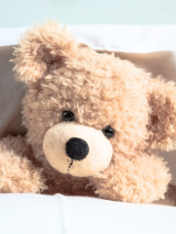 Pluszowy, jasnobeżowy miś leży pod kołdrą, oparty o białą poduszkę