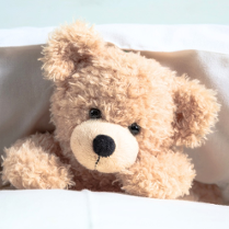 Pluszowy, jasnobeżowy miś leży pod kołdrą, oparty o białą poduszkę