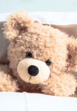 Pluszowy, jasnobeżowy miś leży pod kołdrą, oparty o białą poduszkę