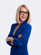 Uśmiechnięta kobieta w okularach z założonymi rękami w niebieskiej marynarce na jednolitym, białym tle.