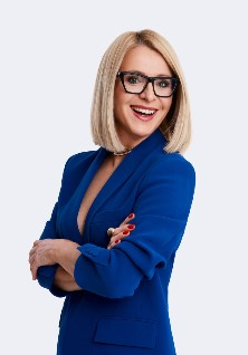 Uśmiechnięta kobieta w okularach z założonymi rękami w niebieskiej marynarce na jednolitym, białym tle.