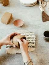 Osoba formuje naczynie z gliny na stole roboczym, na którym leżą różne narzędzia i kawałki gliny.